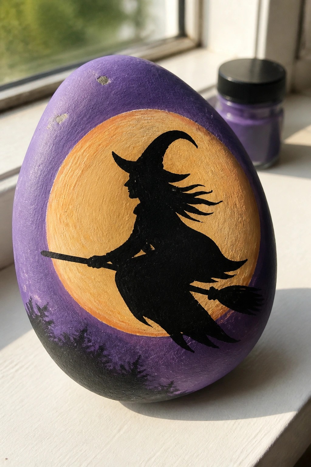 Purple painted rock with black witch silhouette on broomstick before orange moon