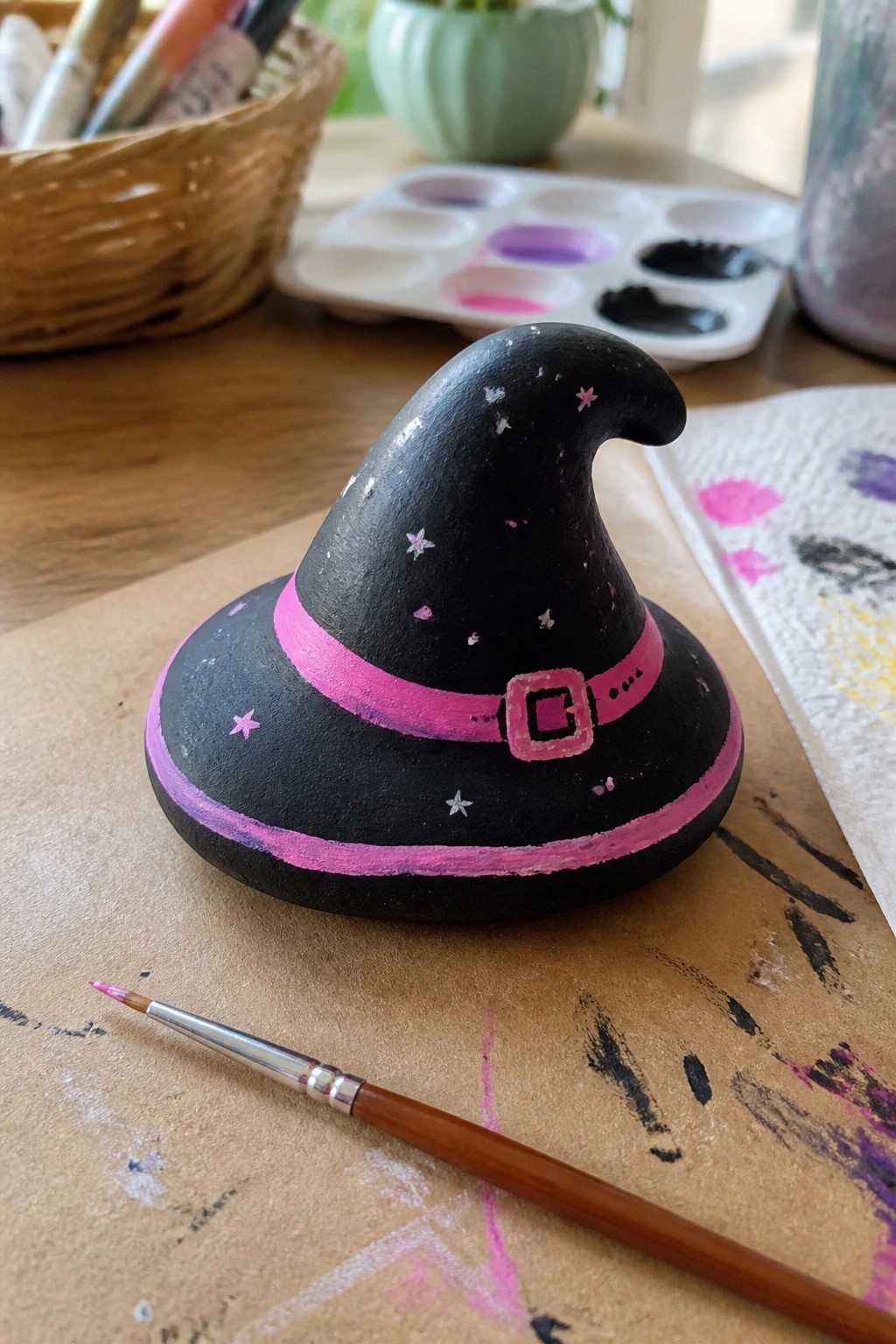 Black painted rock witch hat with pink band, buckle, and stars beside paintbrush