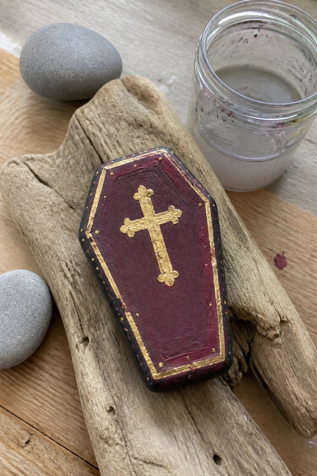 Maroon coffin-shaped box with gold cross on driftwood near stones and jar