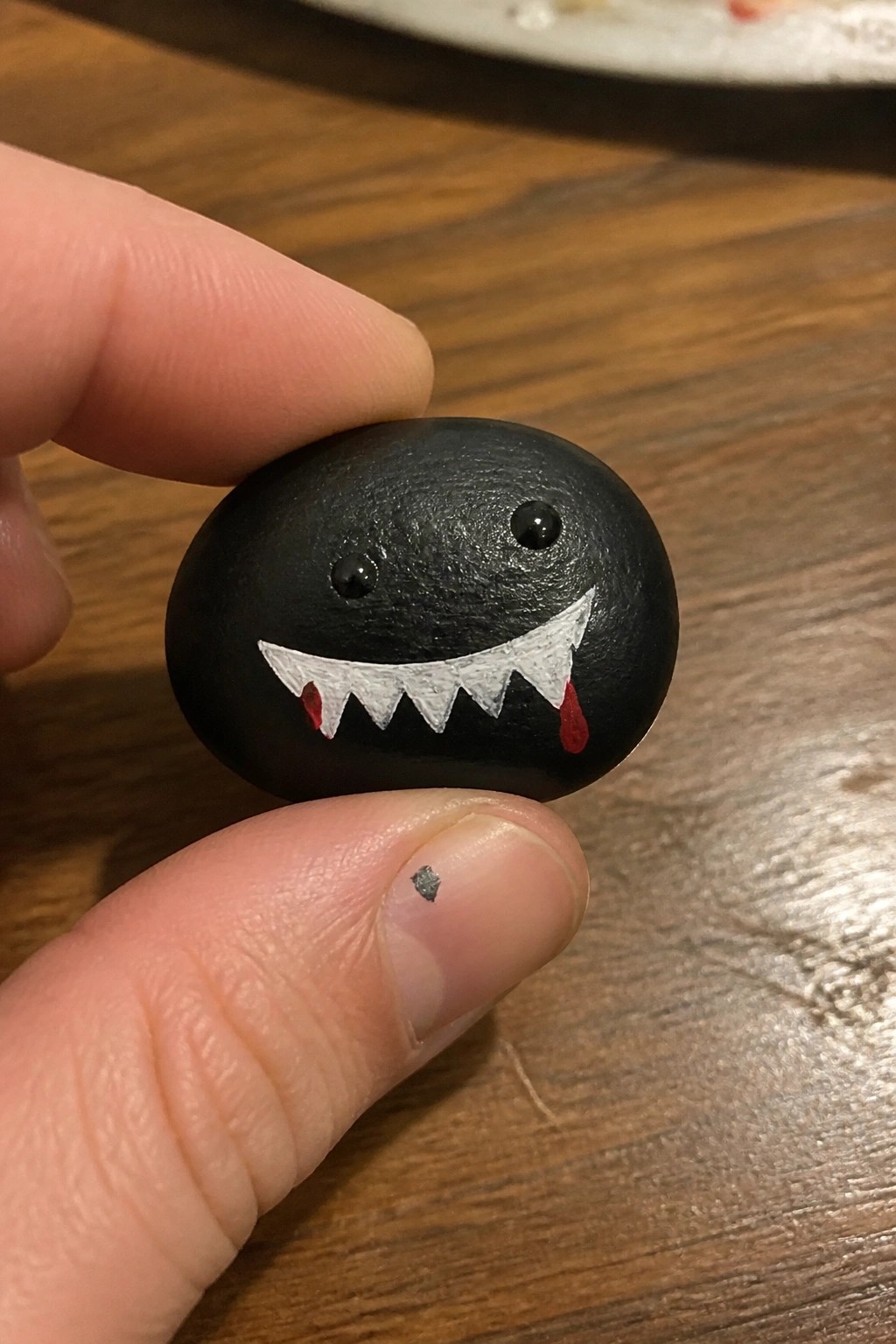 Fingers holding black pebble painted with white jagged teeth and red drips.