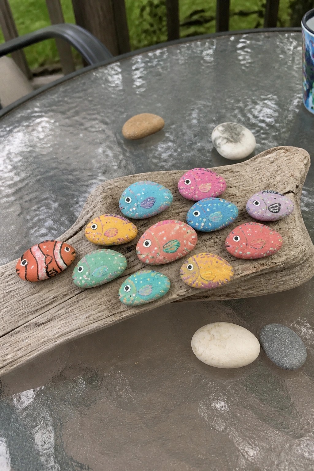 A group of smooth river rocks painted as colorful tropical fish arranged on a piece of driftwood with a few unpainted stones nearby.