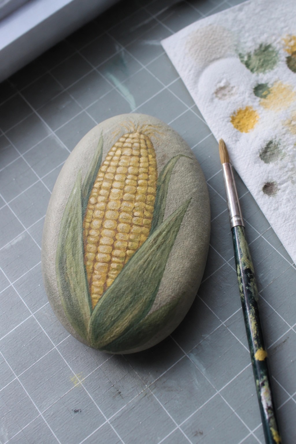 Painted stone corn cob with yellow kernels and green husks beside paintbrush on mat