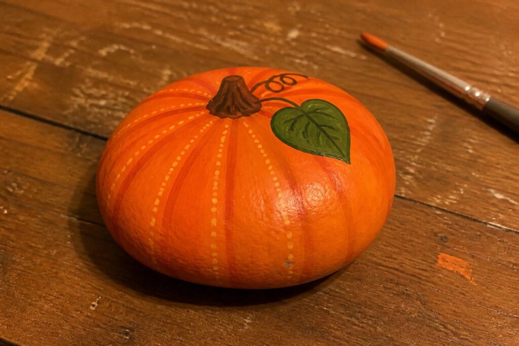 18 Charming Thanksgiving Rock Painting Ideas Centered Around Thankful Designs Painted orange pumpkin with green leaf on wood beside paintbrush and pigment