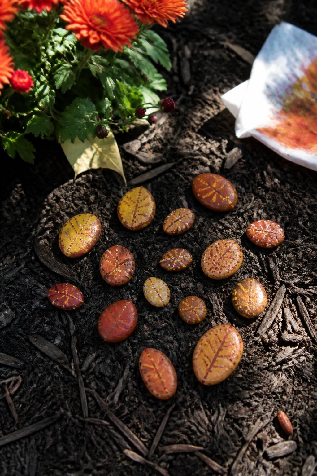 Painted oval stones with leaf vein designs in autumn colors rest on dark mulch near orange chrysanthemums.
