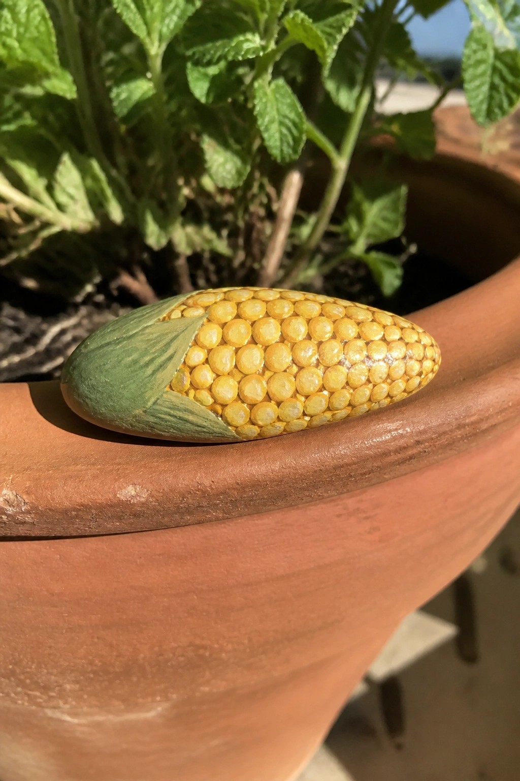 A painted rock resembling a corn cob rests on the rim of a terracotta pot surrounded by green plants.