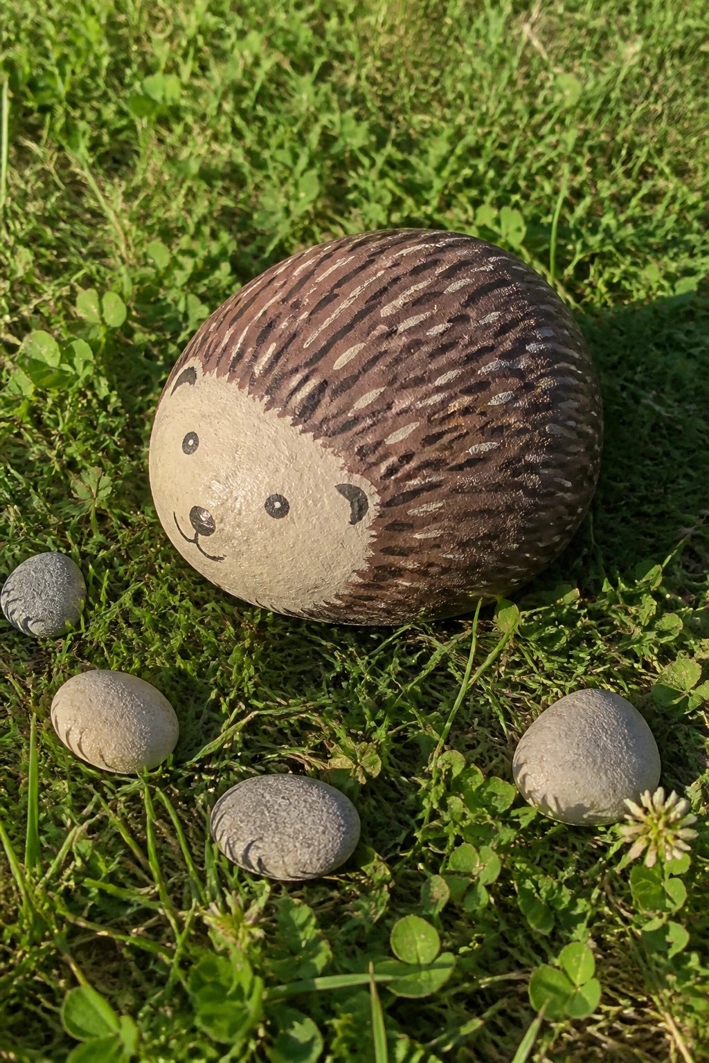 Large hedgehog-painted rock on grass surrounded by smaller stones and clover