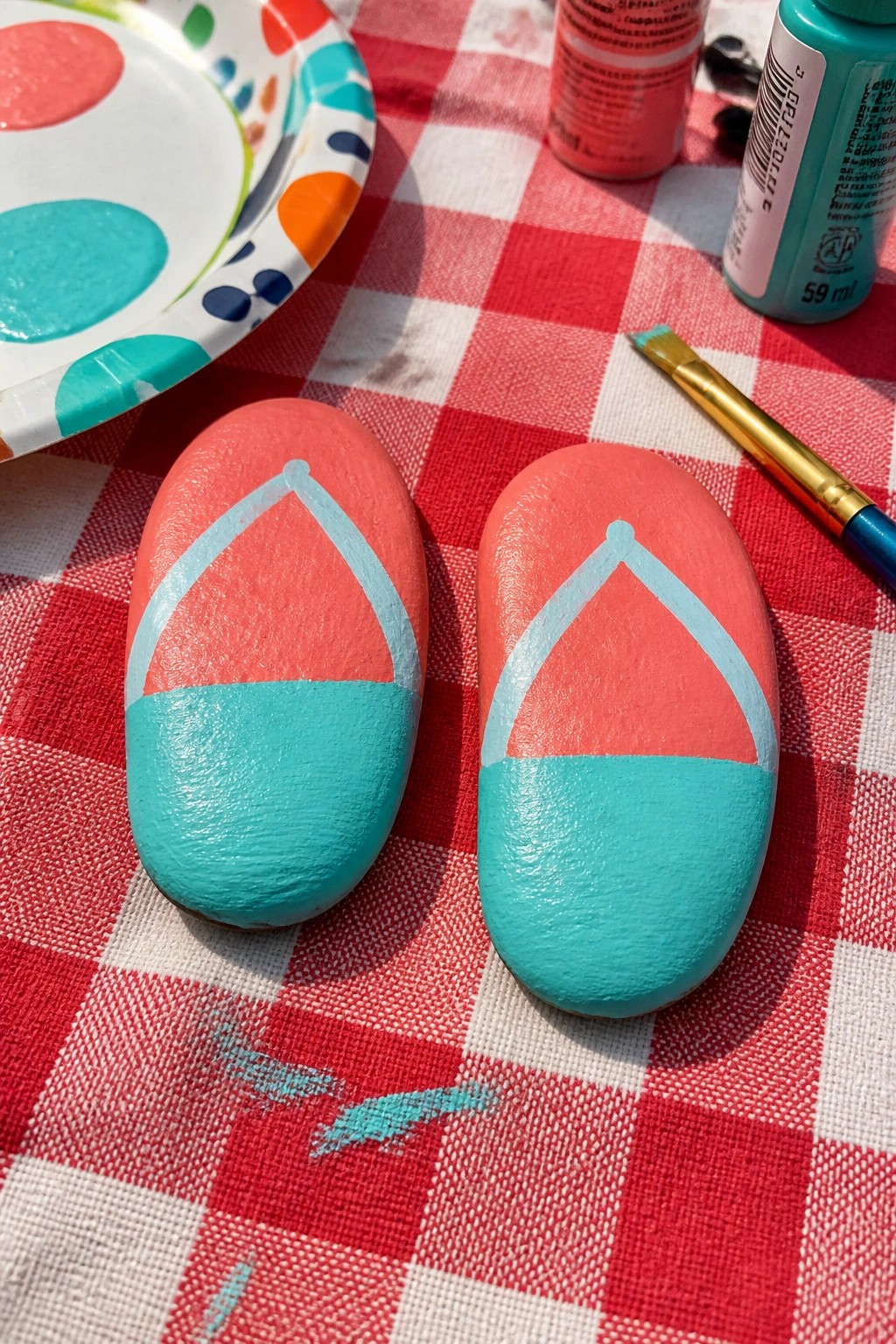 Two rocks painted as coral-turquoise flip-flops on red checkered cloth with paints and brush.