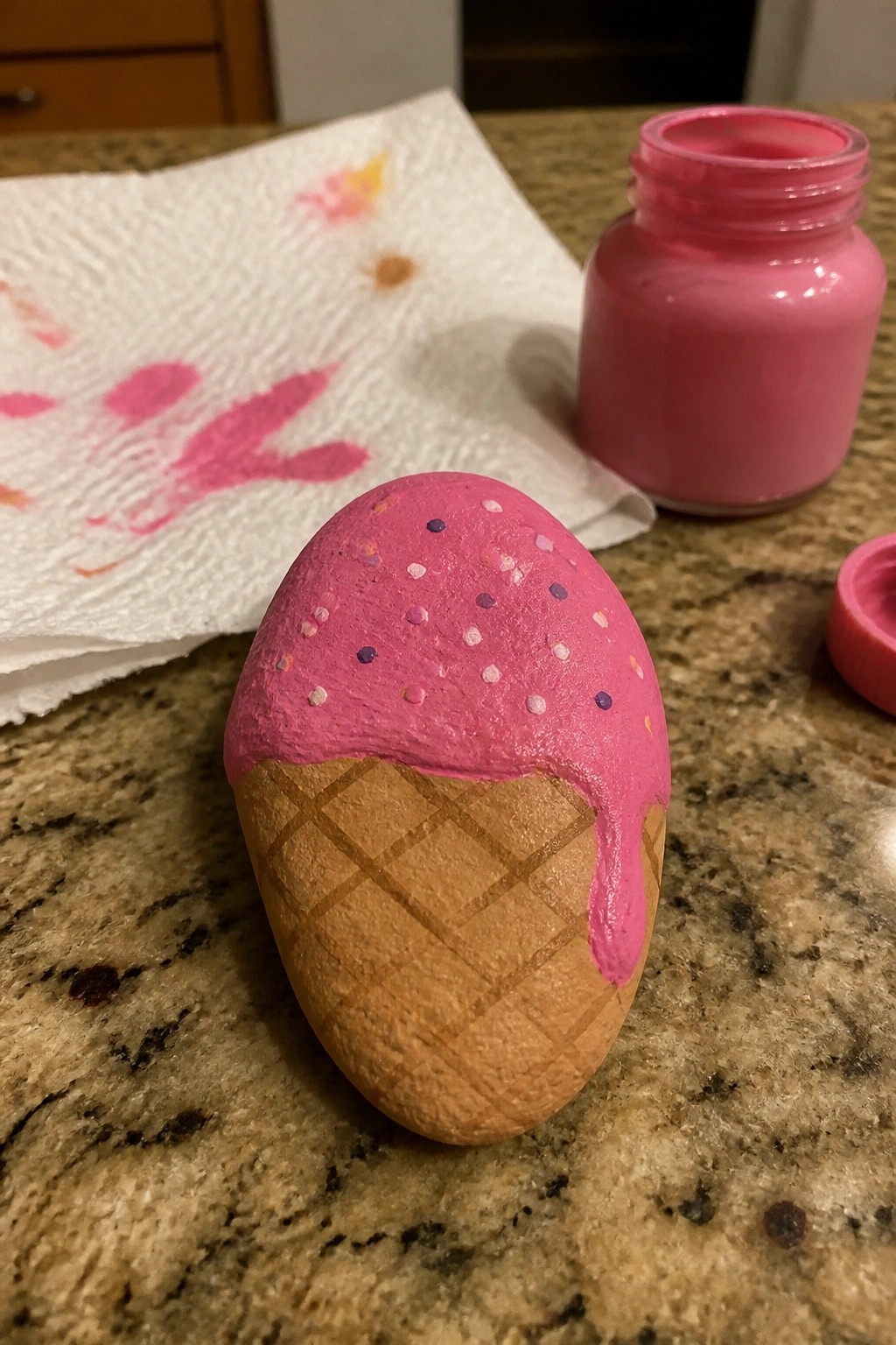 A painted rock resembling an ice cream cone with pink topping and sprinkles sits on a countertop next to a jar of pink paint.
