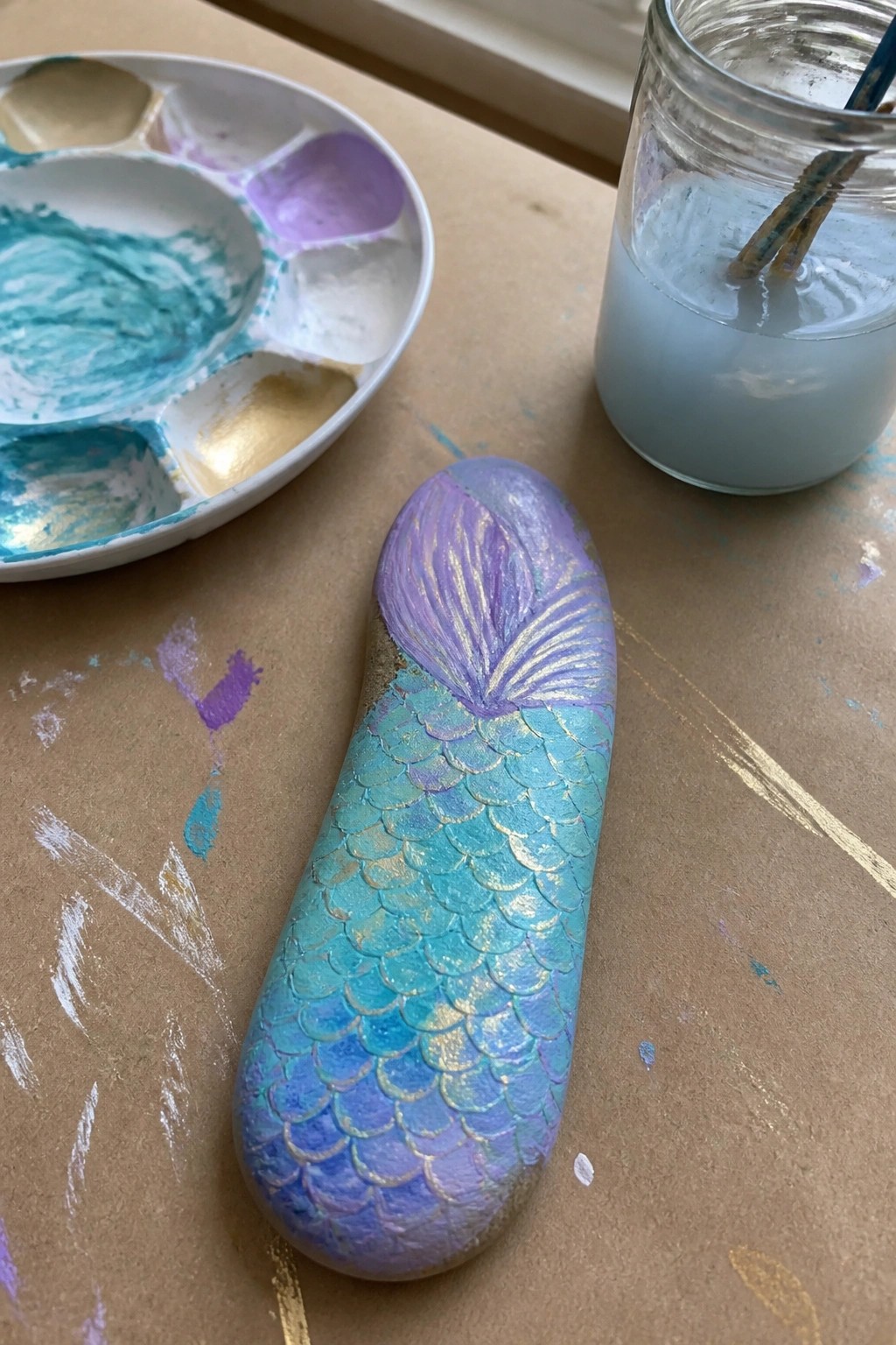 Mermaid tail rock with blue-purple scales beside paint palette and brush jar