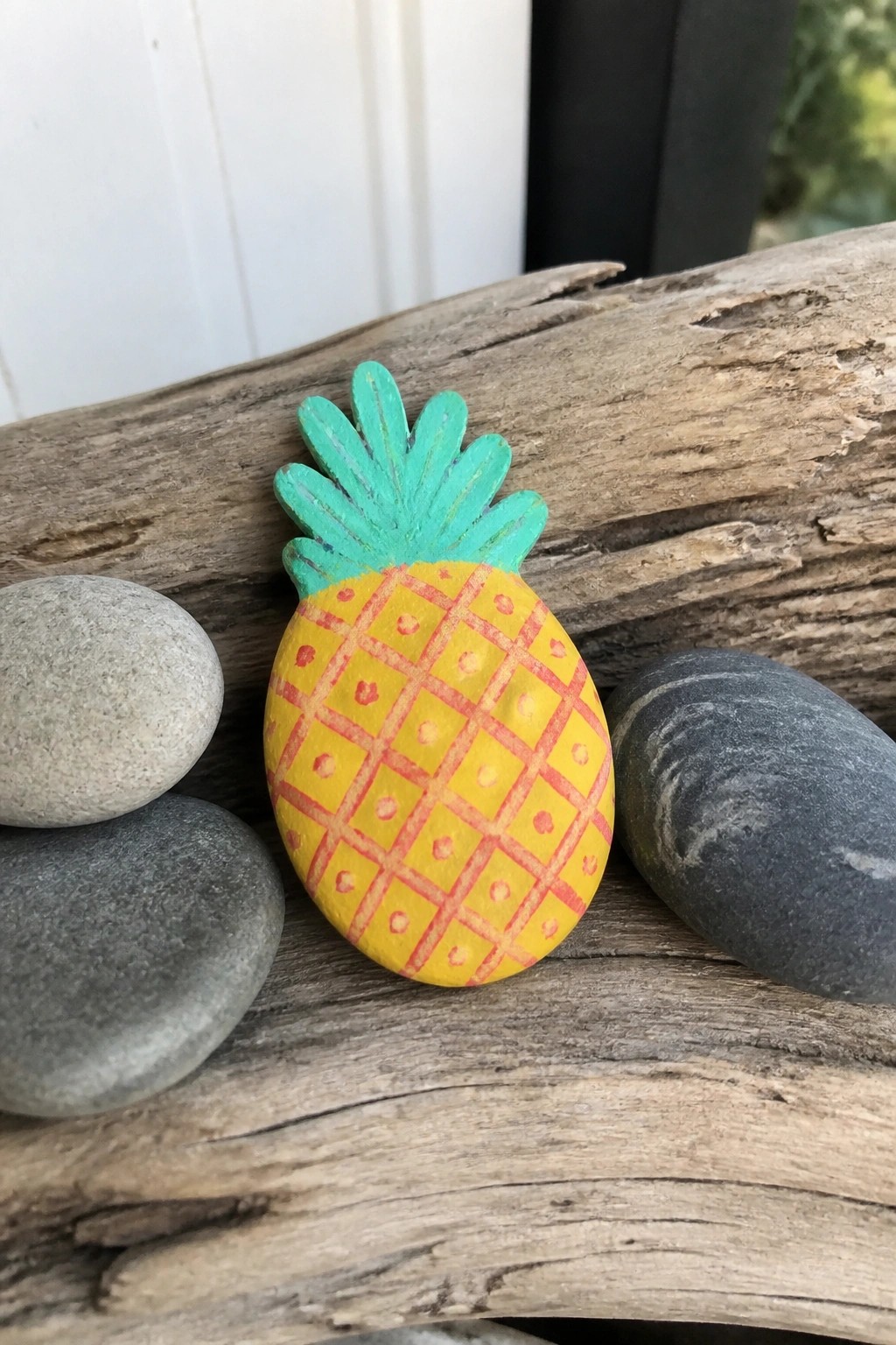 Hand-painted wooden pineapple with yellow body and teal top on driftwood amid stones.