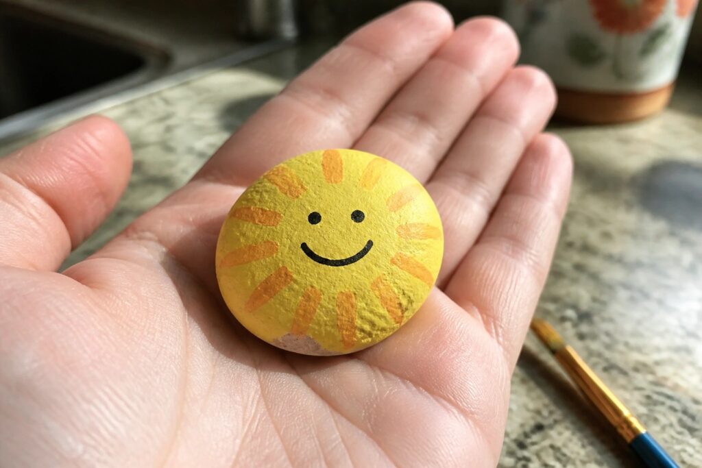 19 Clever Small Rock Painting Ideas Designed for First-Time Painters Hand holding yellow painted rock with smiling sun face and orange rays.