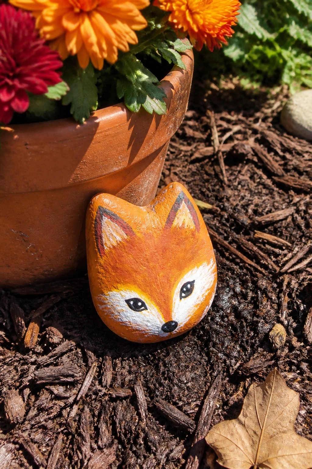A small rounded rock painted orange and white as a fox face sits on dark mulch beside a terracotta flower pot.