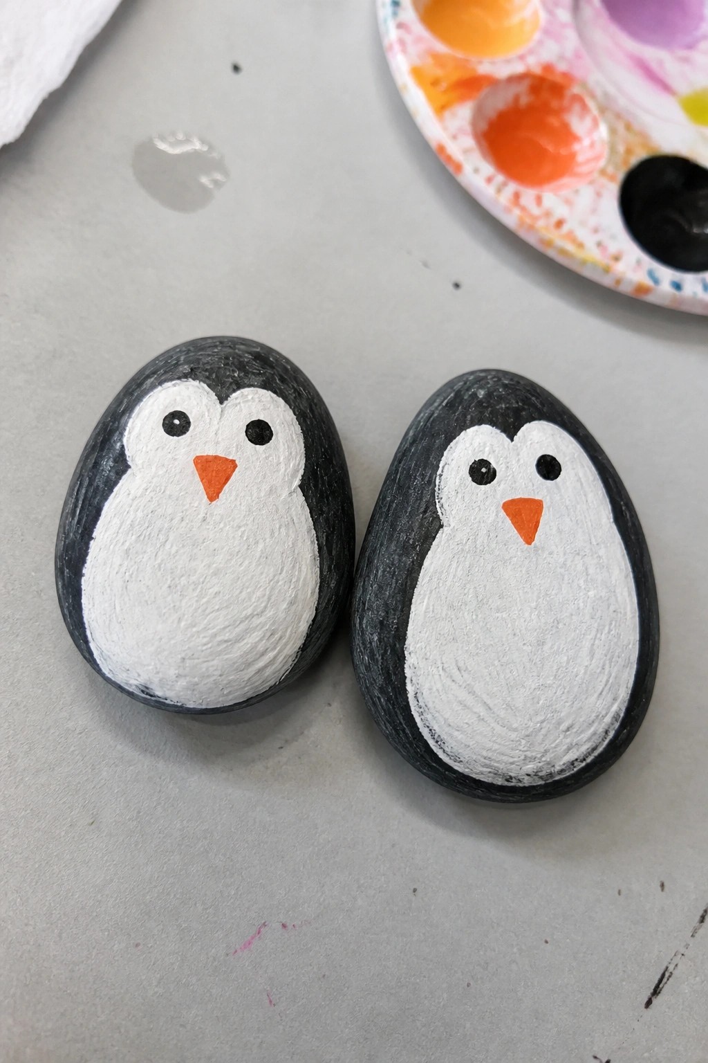 Two hand-painted rocks as penguins with orange beaks beside a palette.