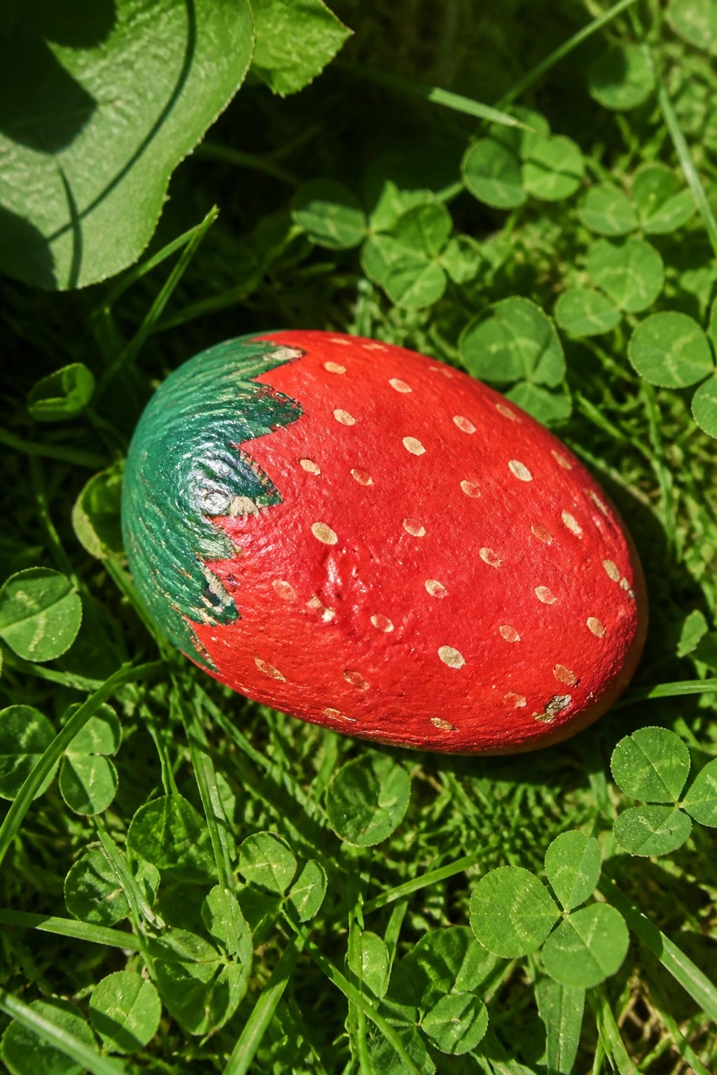 Red strawberry-shaped painted rock with white dots on green clover grass.