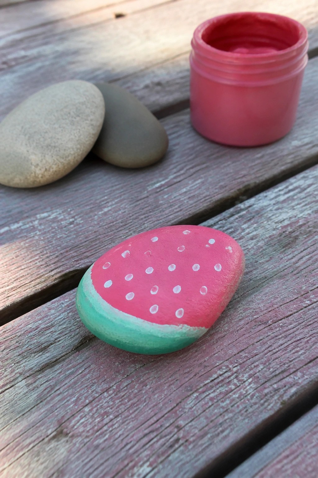 Watermelon-painted rock beside open pink paint jar on wooden table