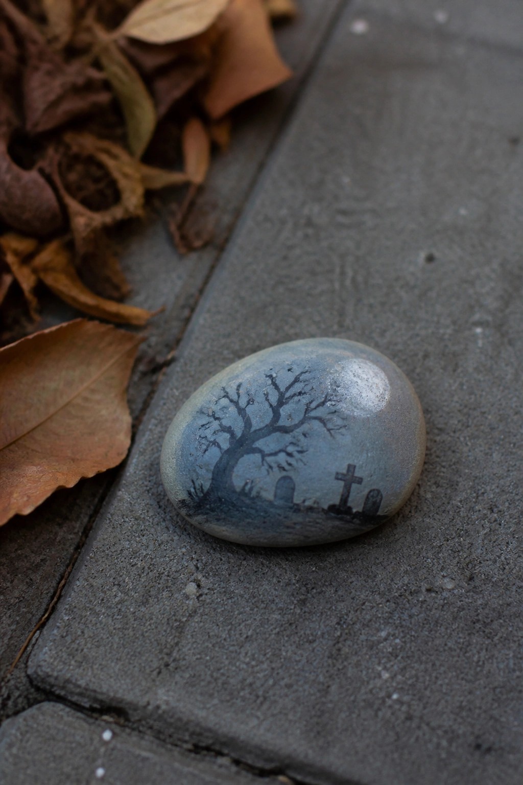 Painted stone showing bare tree and graveyard on gray pavement near dry leaves