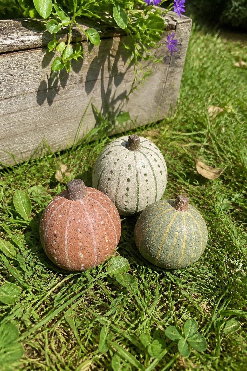 Three painted rocks resembling small pumpkins rest on grass next to a wooden planter.