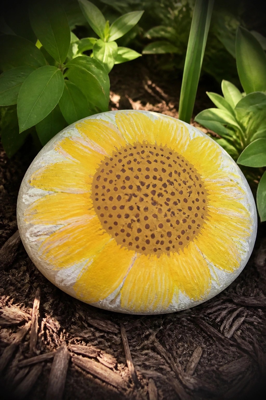 A round stone painted with a yellow sunflower and brown dotted center sits on dark mulch near green leaves.