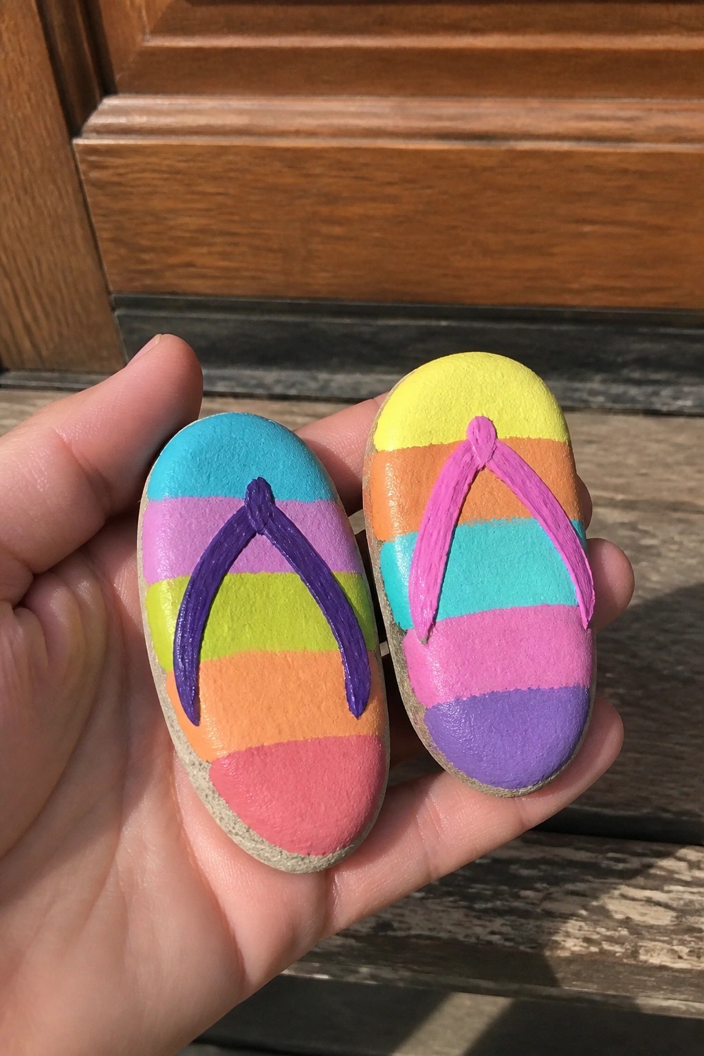 Hand holding two rainbow-striped rocks painted as flip-flop sandals with straps