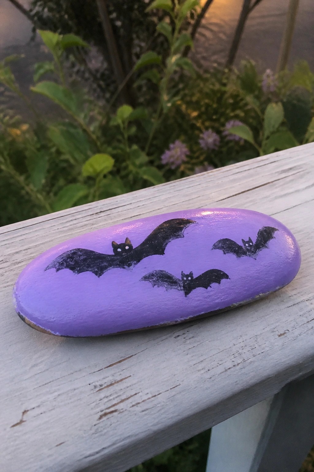 A purple painted oval rock with three black bats sits on a white wooden railing.