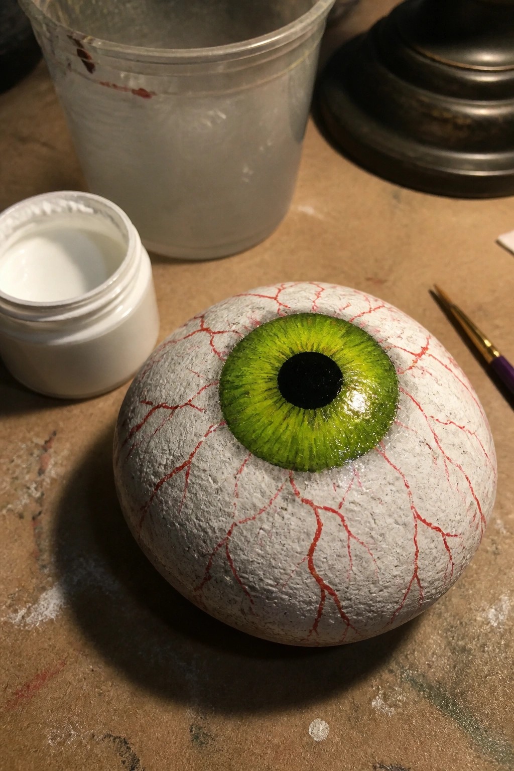 Painted green eyeball with red veins beside white paint jar and brush