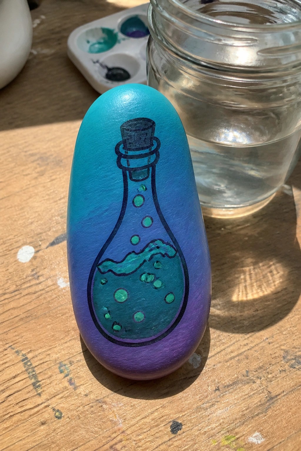 Oval painted rock with blue-purple gradient and illustrated green bubbling potion bottle