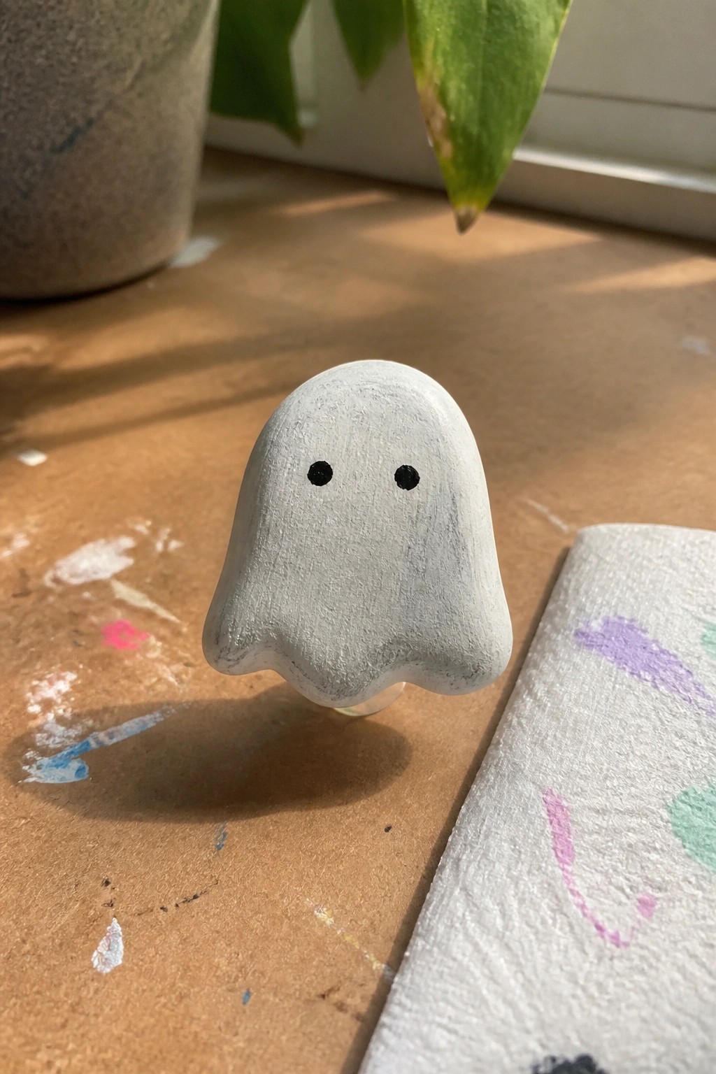 Small white clay ghost figurine with black eyes on paint-stained wooden table