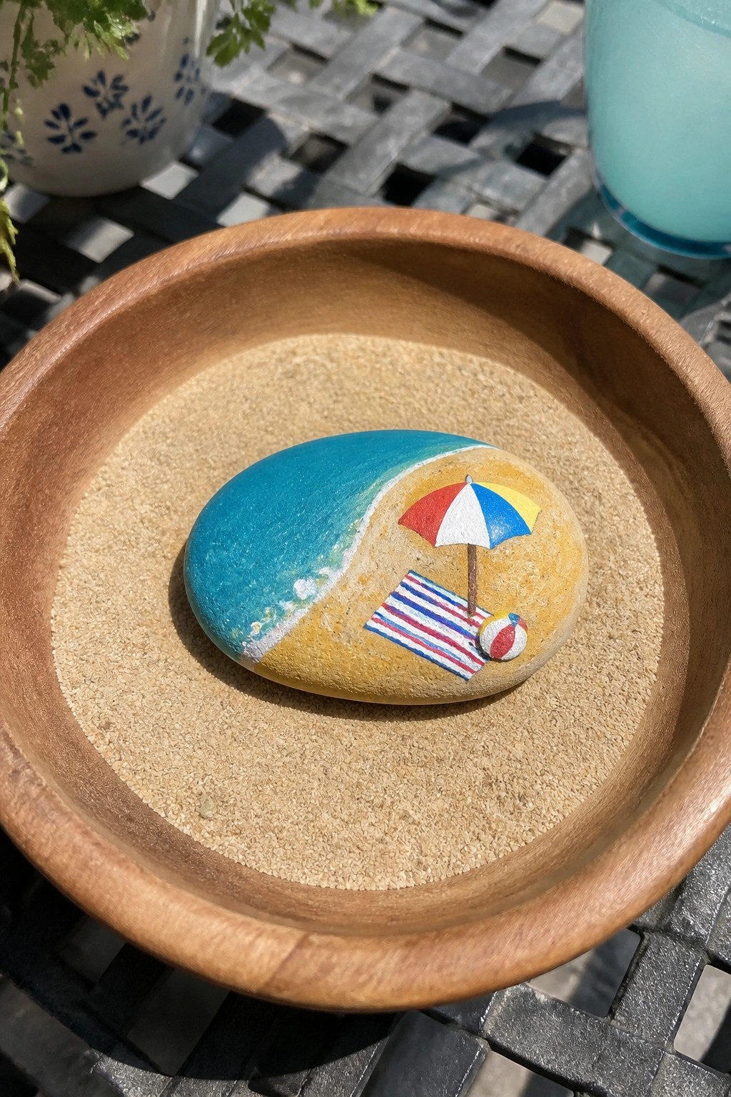 A painted oval rock with blue water, yellow sand, umbrella, towel, and ball rests in a sand-filled wooden bowl.