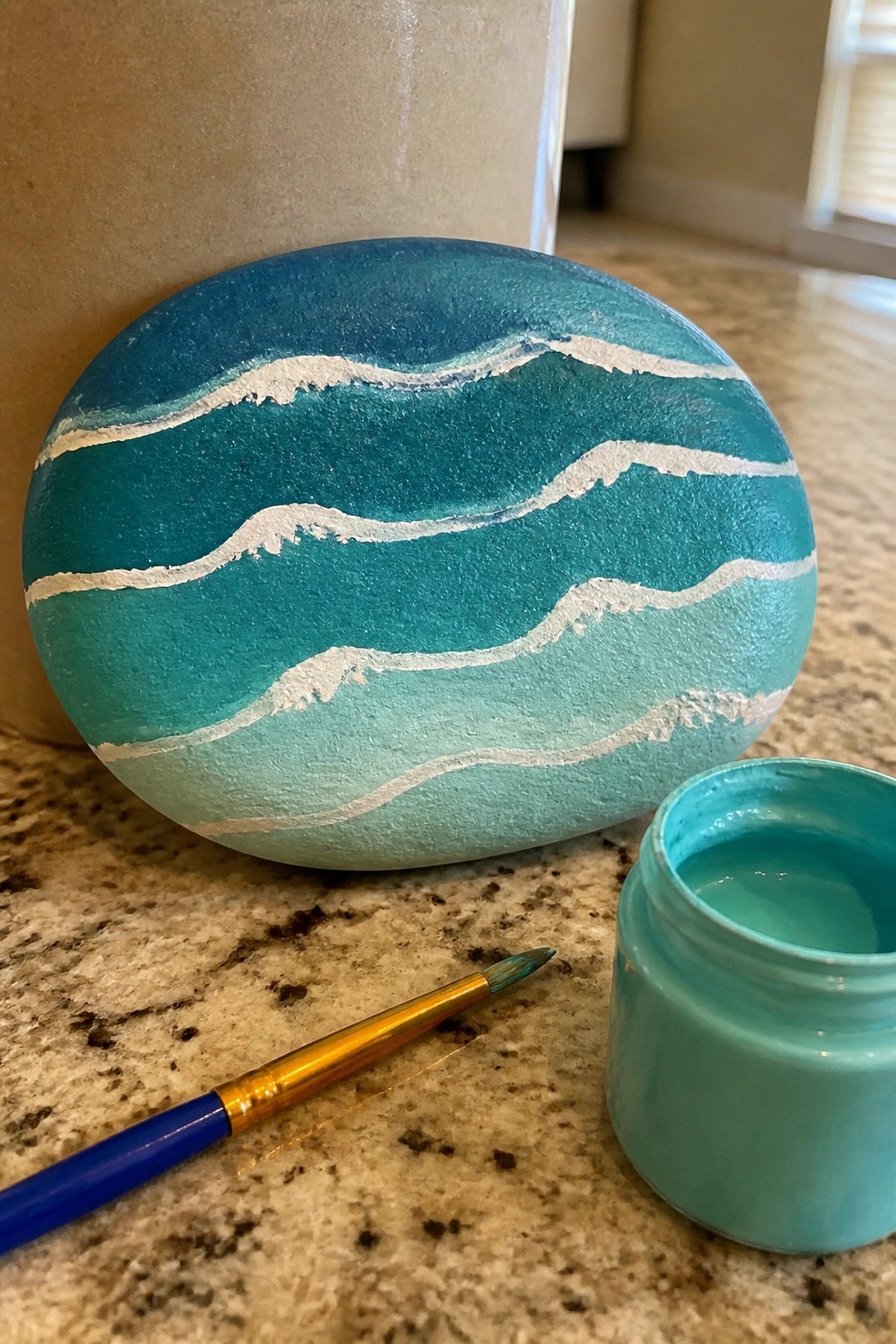 A smooth oval rock painted with blue and teal ocean waves sits on a countertop next to a paint jar and brush.