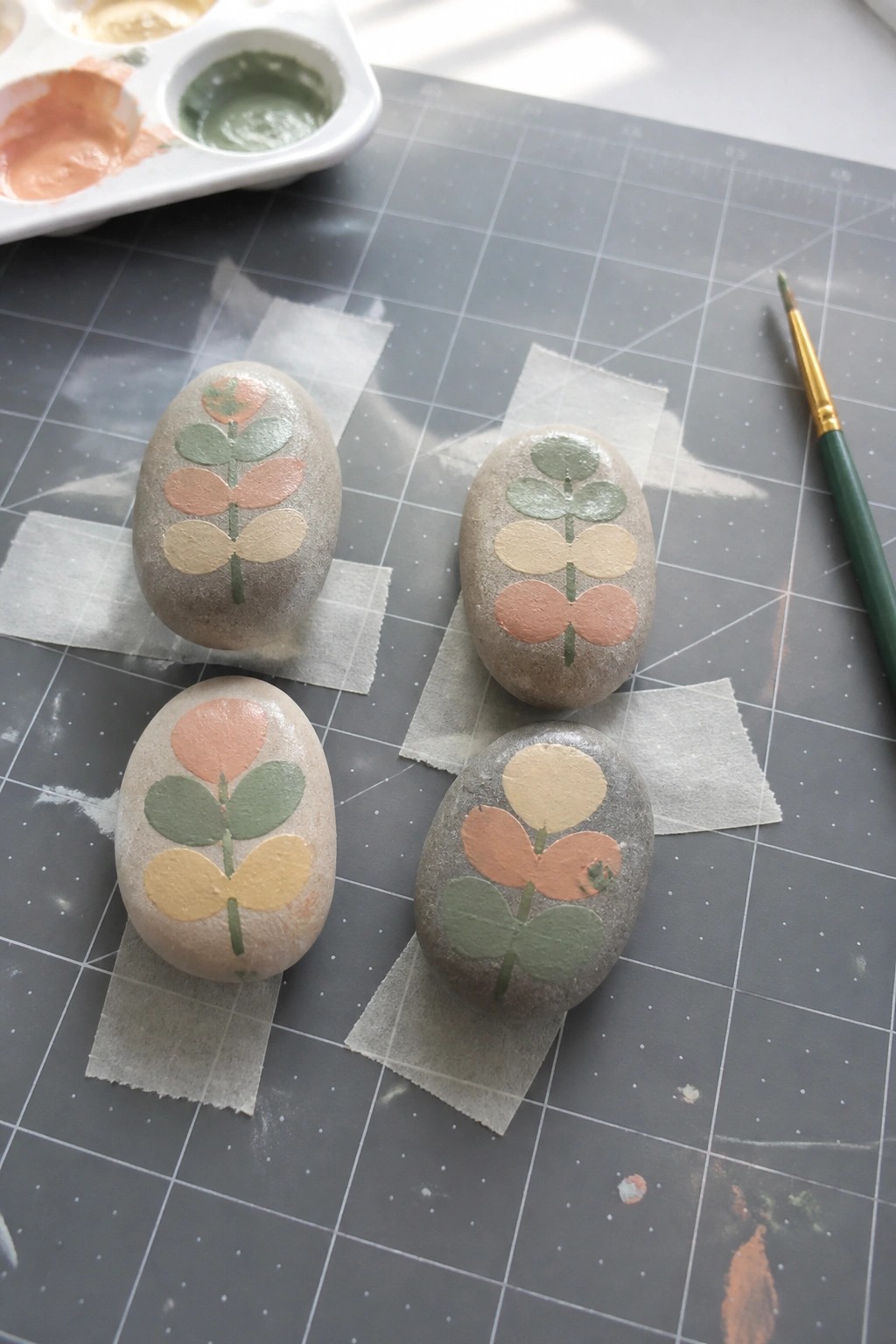 Four painted rocks with floral designs on a gray mat beside paints and brush.