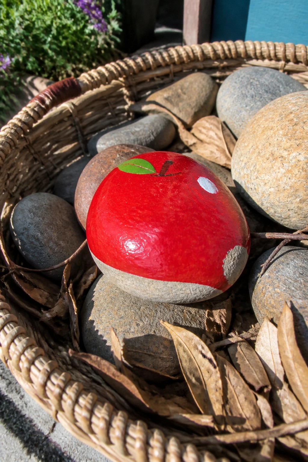 Red apple rock with green leaf in wicker basket amid stones and leaves.