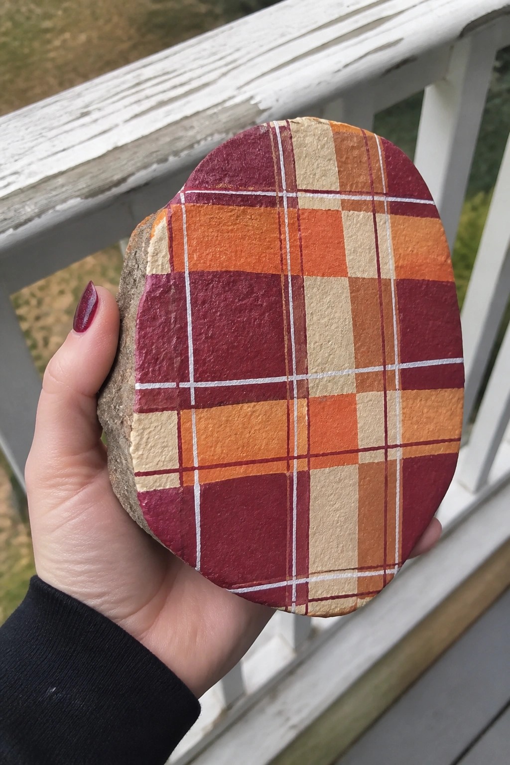 Hand holding oval rock painted with maroon-orange plaid pattern on porch.