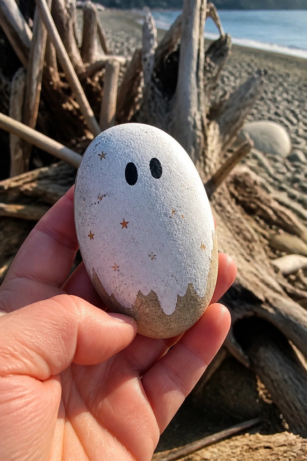 Hand holding white ghost-painted rock with black eyes and gold stars on beach