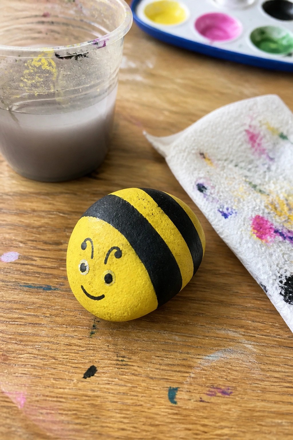 A yellow and black striped bee painted on a rounded rock with a simple face sits on a wooden table next to a cup of water and a paint palette.