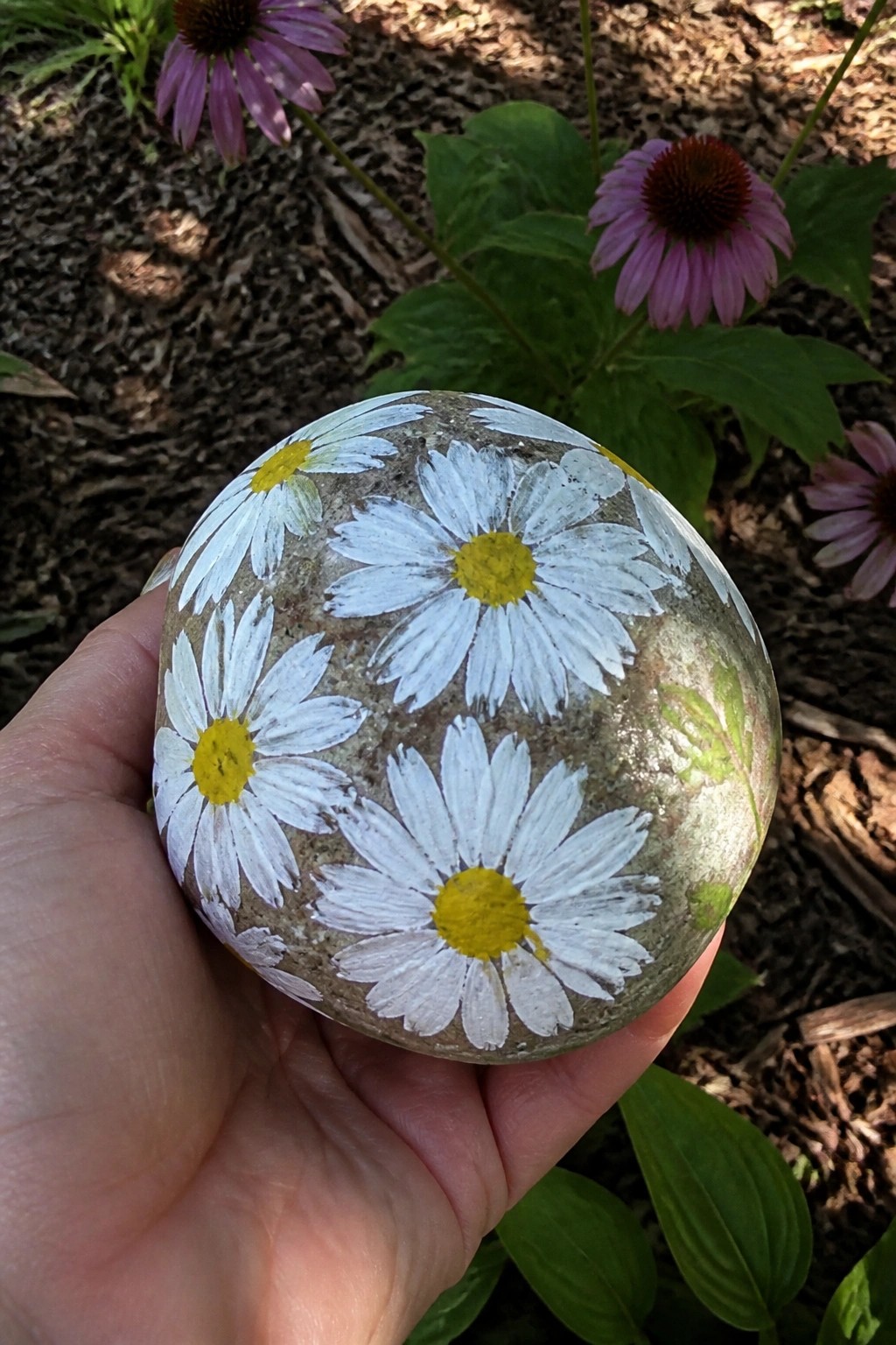 Hand holding rock painted with white daisies and yellow centers outdoors