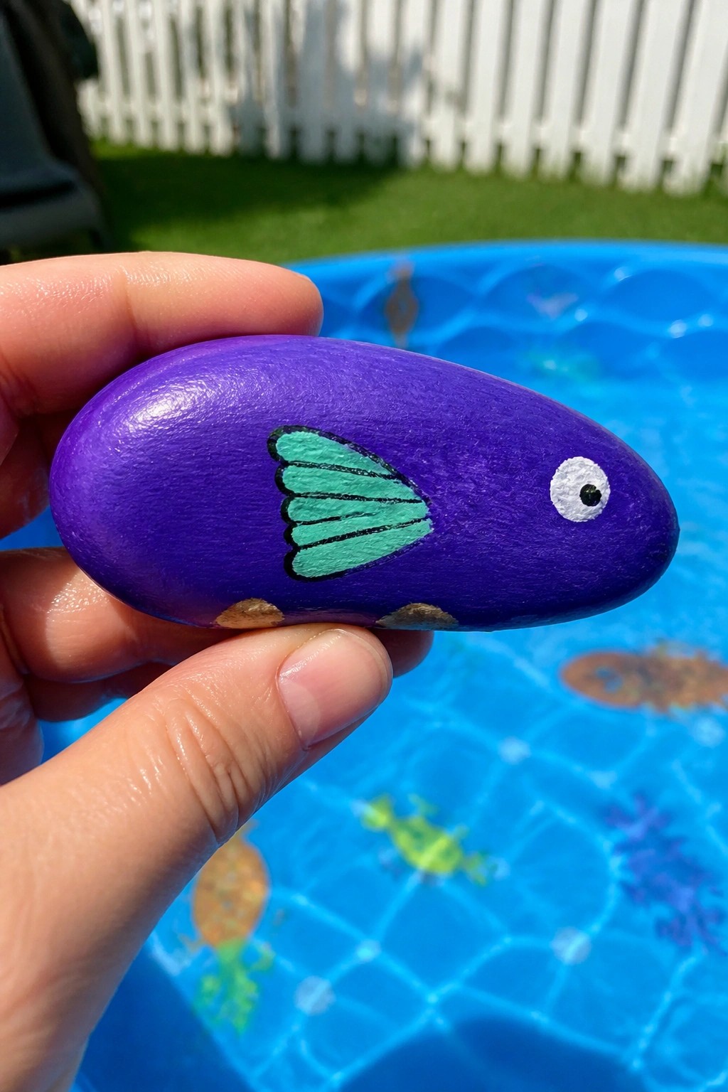 Hand holding glossy purple fish rock with green fin over blue pool