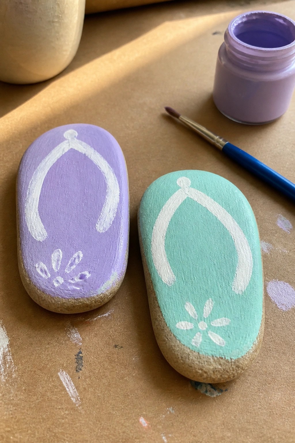 Two oval rocks painted purple and mint green with white flip-flop strap outlines and flower details sit on brown paper next to a jar of purple paint and a brush.