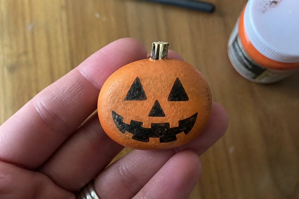 21 Clever Pumpkin Rock Painting Ideas to Celebrate the Halloween Season Hand holding orange rock painted as jack-o'-lantern beside paint bottle on wood