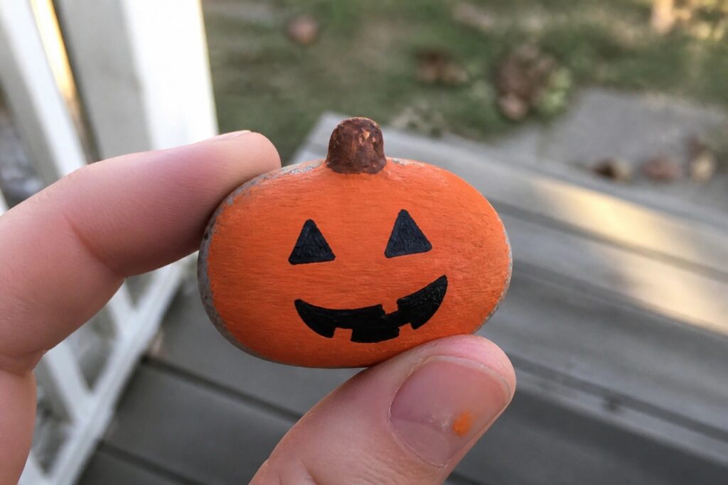 21 Creative Halloween Pumpkin Painted Rock Ideas Without Complicated Designs Fingers holding small orange pumpkin with black jack-o'-lantern face on wooden deck.