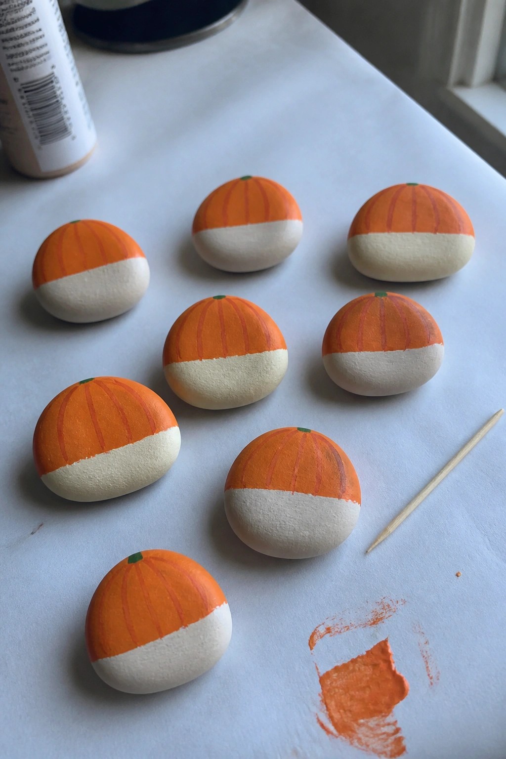 Eight painted pumpkin rocks on white table with orange paint smear and skewer