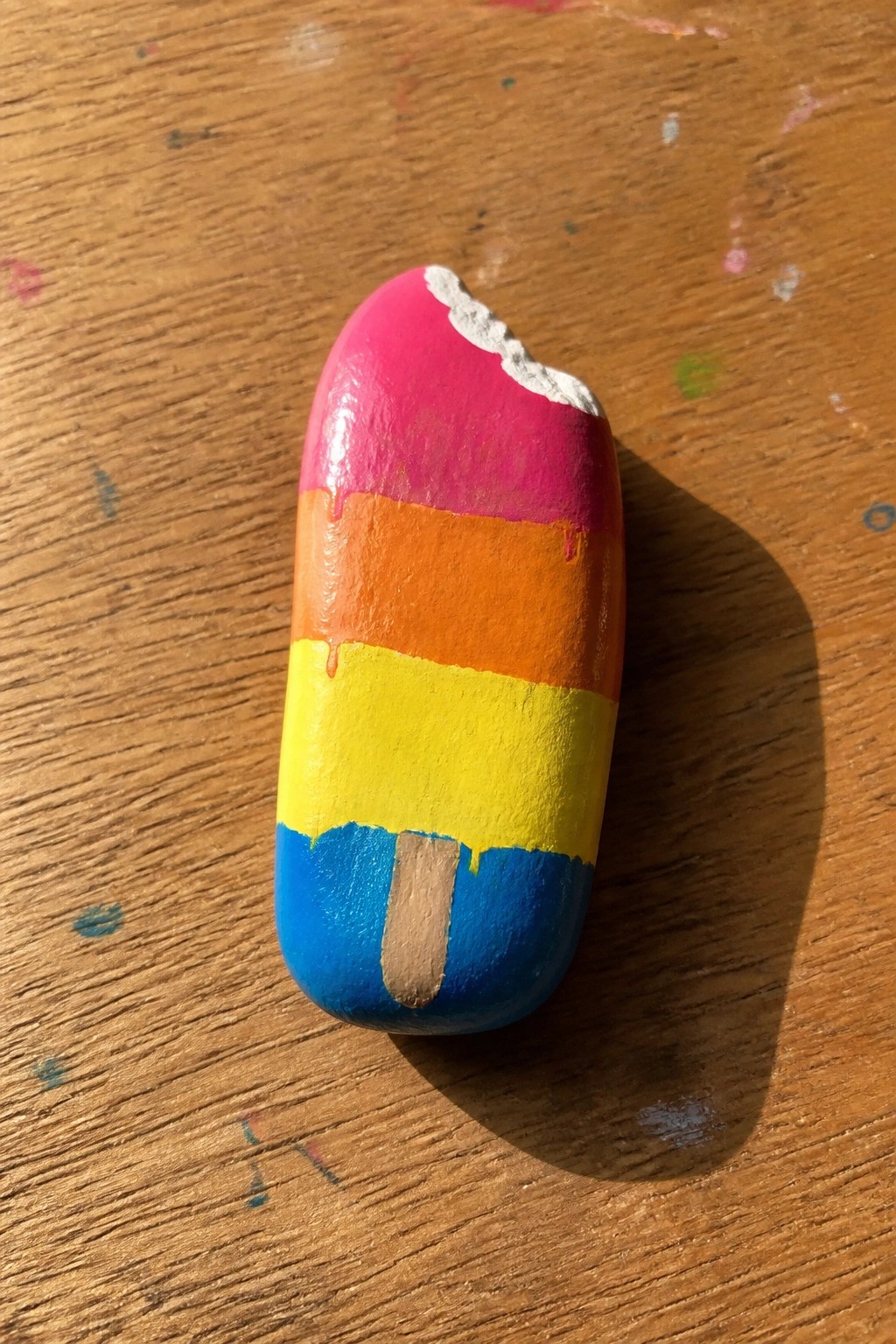 Painted rock shaped like a rainbow popsicle with pink, orange, yellow, blue stripes and stick