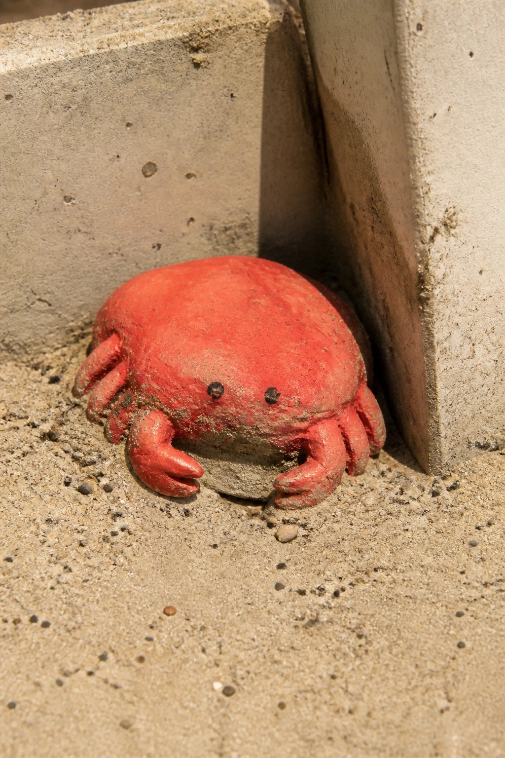 A red painted rock designed as a crab rests on sandy ground next to a concrete surface.