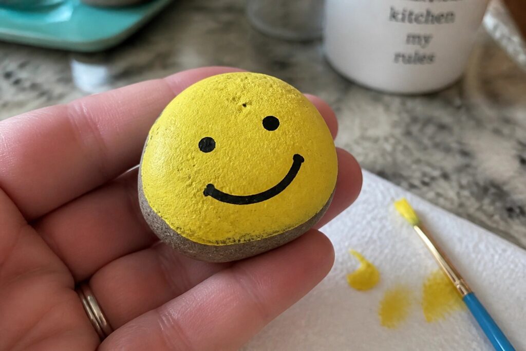 21 Easy Summer Rock Painting Ideas for Beginners Using Simple Shapes and Patterns Hand holding small yellow smiley face painted on round stone