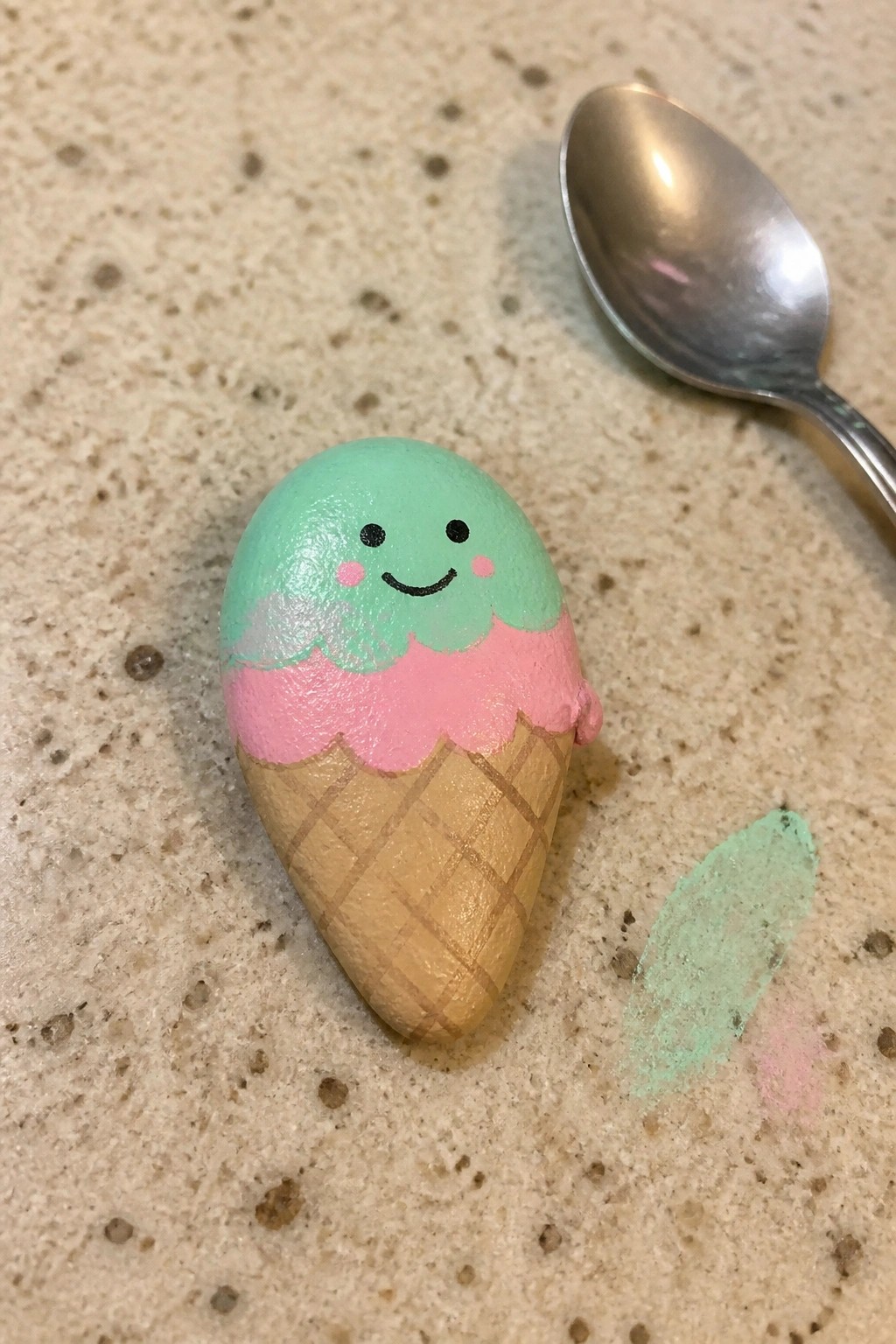 Painted rock ice cream cone with smiling face beside metal spoon on counter