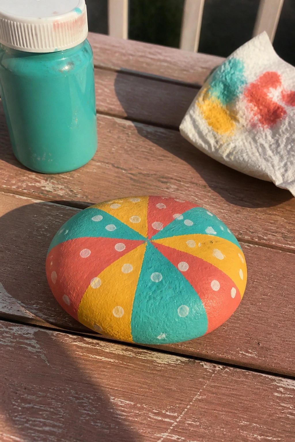 Painted rock with red, yellow, teal wedges and dots beside paint jar on wood