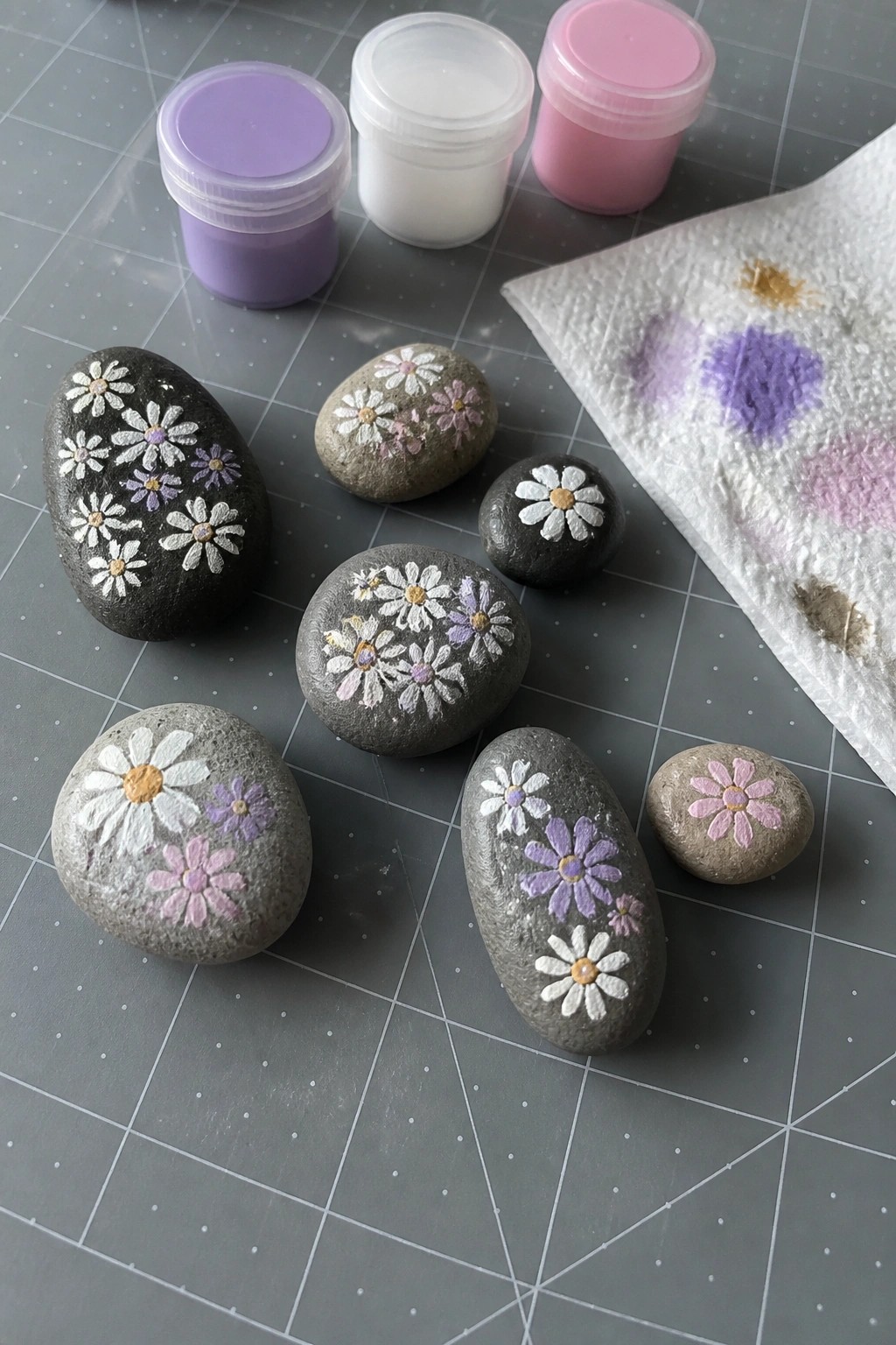 Painted rocks with daisy flowers beside paint jars and stained paper towel on mat.