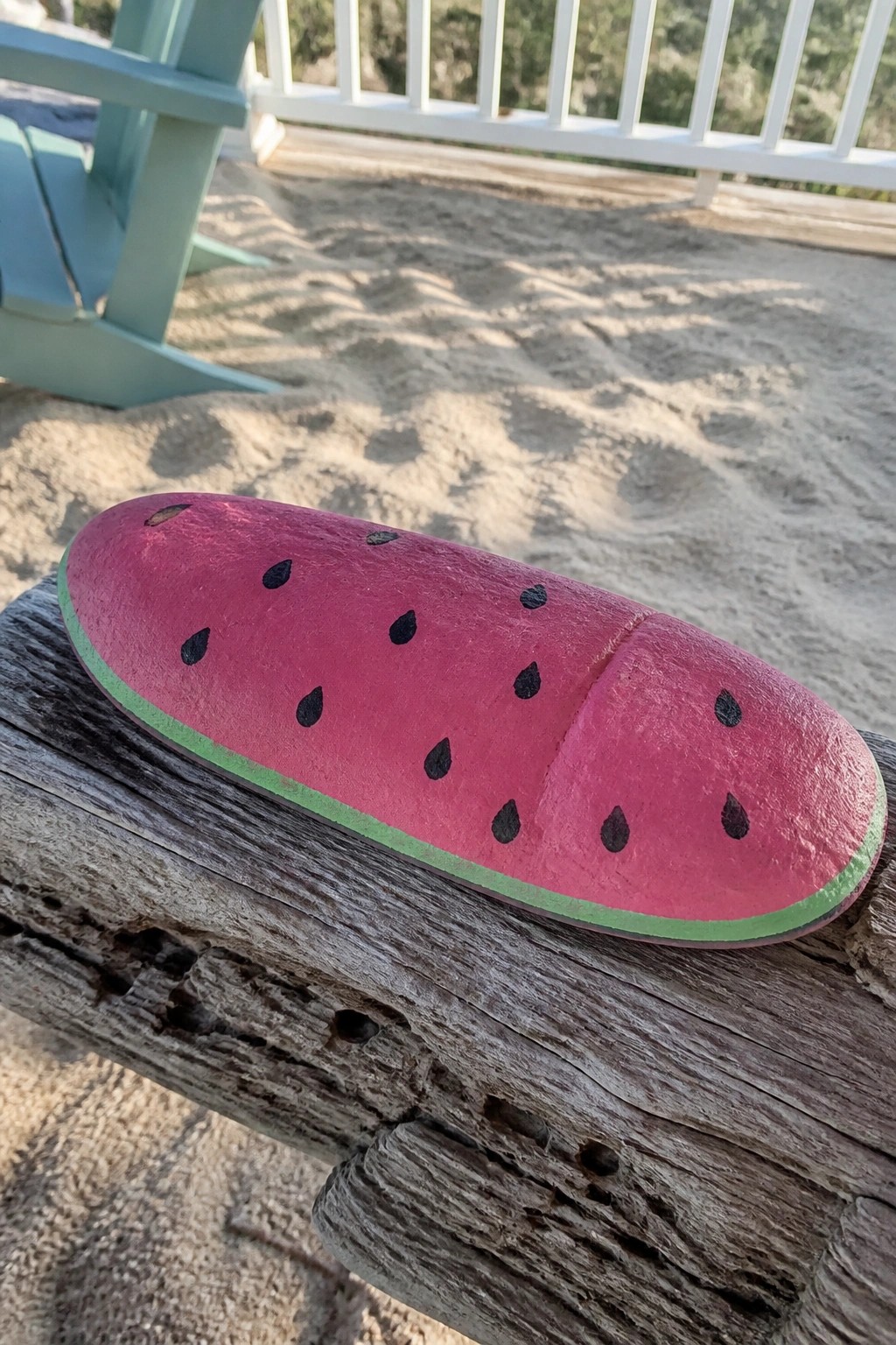 Pink wooden watermelon sculpture with black seeds on weathered beachside bench