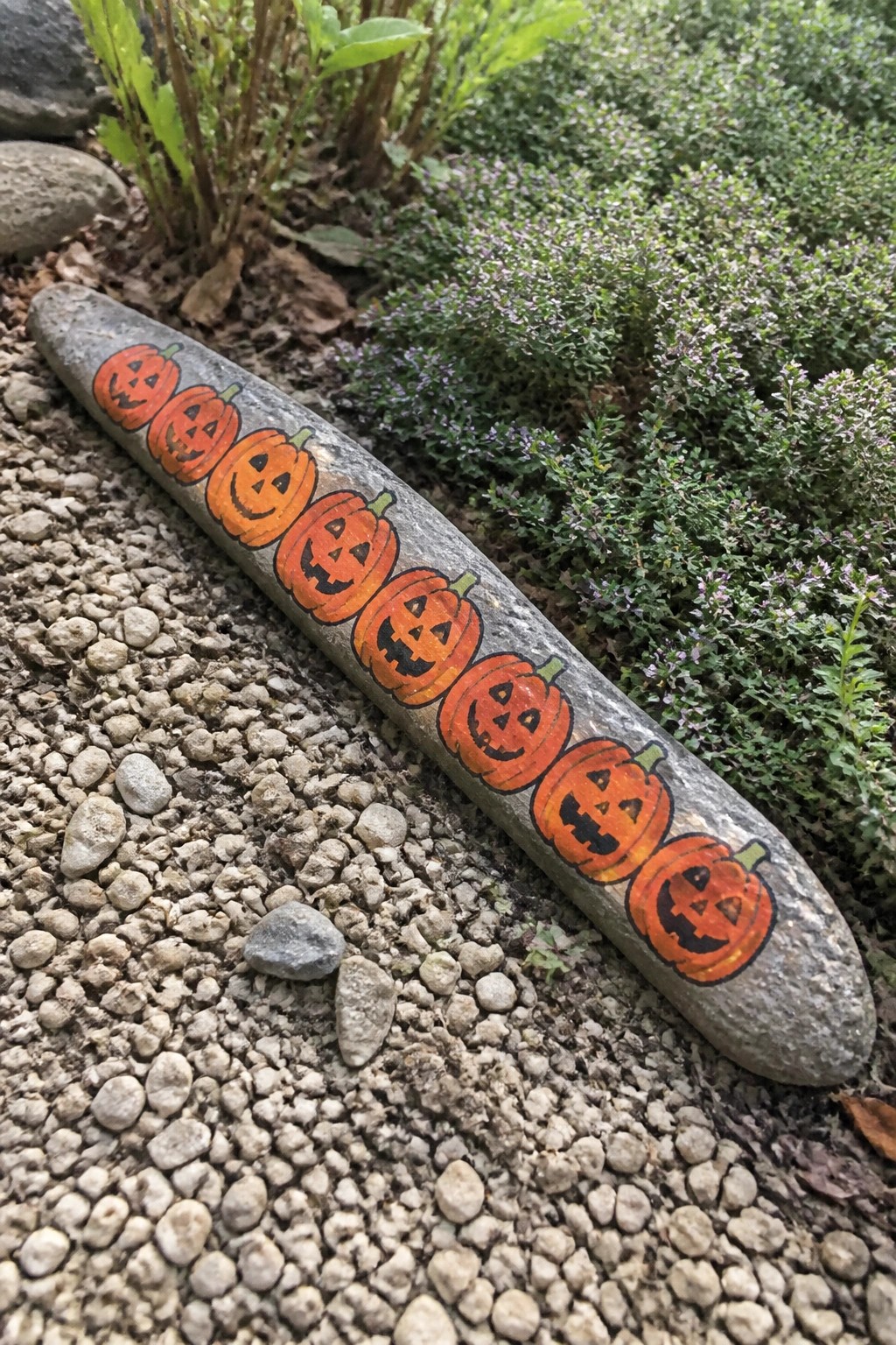 Gray rock painted with row of orange jack-o'-lanterns on gravel ground.