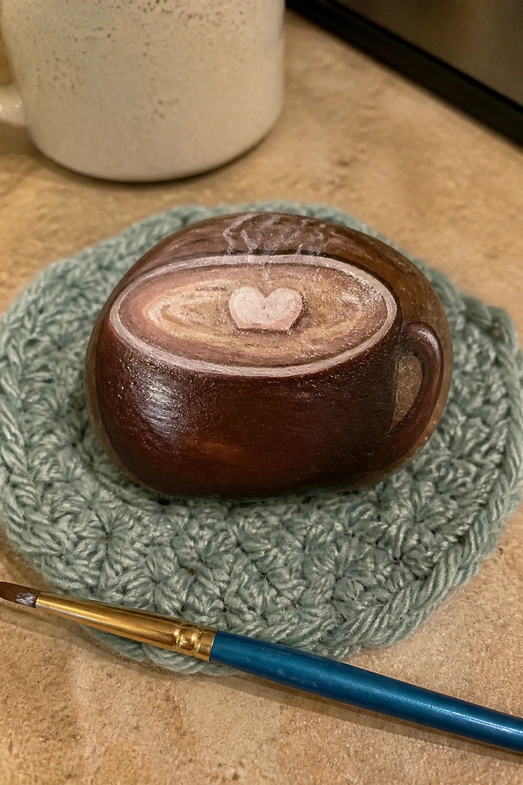 Hand-painted rock coffee cup with heart on green crochet coaster beside paintbrush