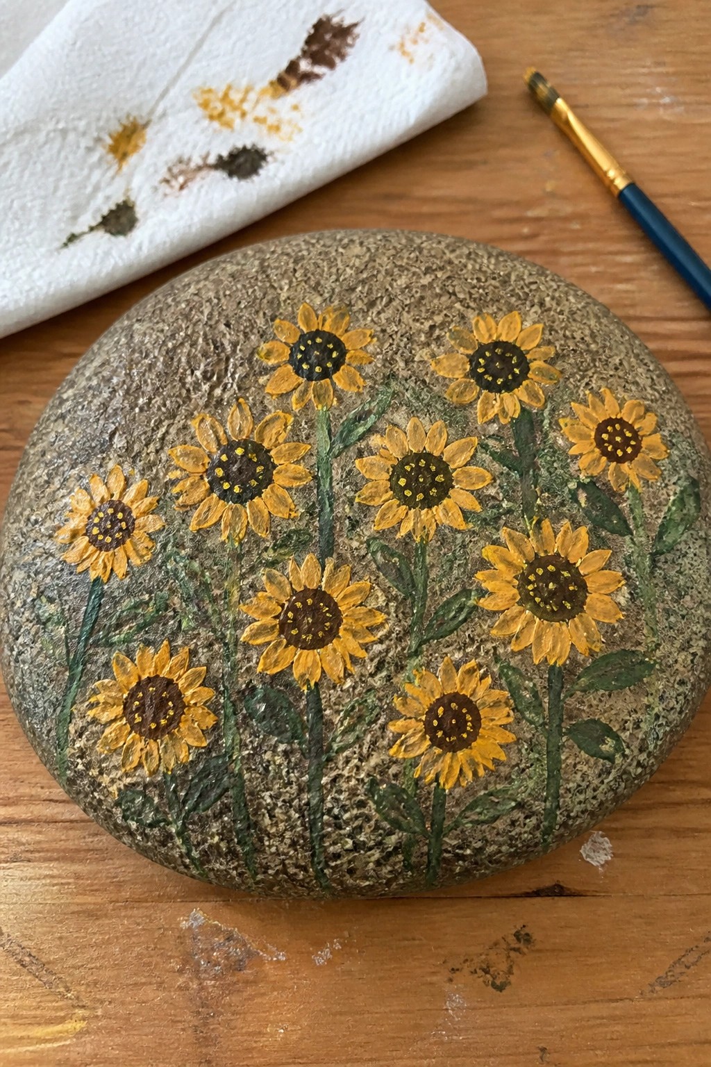 Painted rock with yellow sunflowers and green stems beside brush and stained towel on wood